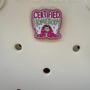 K&ouml;nnte beinhalten: Ein pinkfarbener Glitzer-Anstecker mit den Worten "CERTIFIED HOMEBODY" in Pink und Gr&uuml;n. Der Anstecker zeigt einen Cartoon-Frosch, der in eine rosa Decke mit wei&szlig;en und pinkfarbenen Blumenmustern geh&uuml;llt ist. Auf wei&szlig;em Hintergrund.