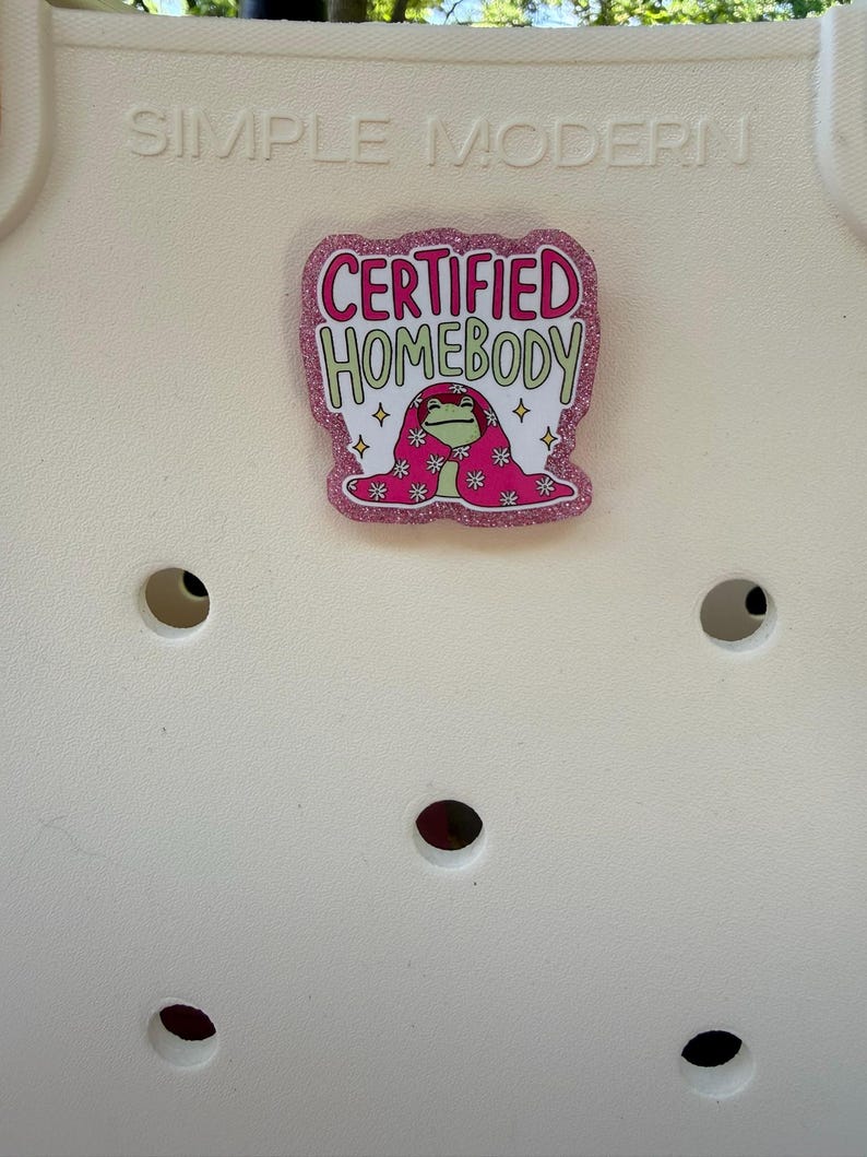 K&ouml;nnte beinhalten: Ein pinkfarbener Glitzer-Anstecker mit den Worten "CERTIFIED HOMEBODY" in Pink und Gr&uuml;n. Der Anstecker zeigt einen Cartoon-Frosch, der in eine rosa Decke mit wei&szlig;en und pinkfarbenen Blumenmustern geh&uuml;llt ist. Auf wei&szlig;em Hintergrund.