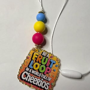 May include: A lanyard with a pendant that reads "Be a Fruit Loop in a World Full of Cheerios." The pendant is rectangular with a gold border and colorful text. The lanyard has three colorful beads and a white cord.