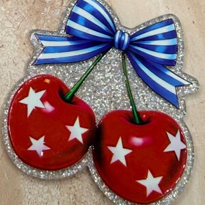 May include: A decorative cherry-shaped ornament featuring two glossy red cherries adorned with white stars. The cherries are connected by green stems topped with a large blue and white striped bow. The entire piece has a silver glitter border.