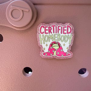 K&ouml;nnte beinhalten: Ein rosa Schuh mit einem dekorativen Charm, auf dem "CERTIFIED HOMEBODY" steht. Der Charm zeigt einen Cartoon-Frosch, der eine rosa Decke mit wei&szlig;en Blumen tr&auml;gt. Der Charm hat einen glitzernden rosa Rand.