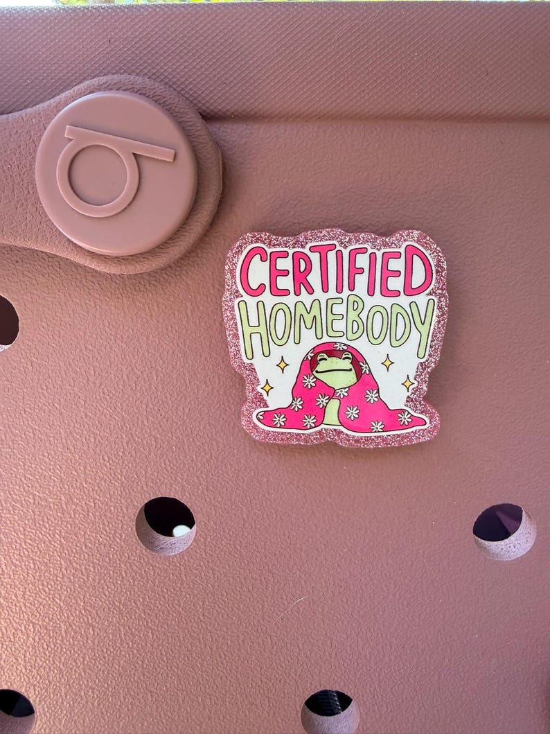 K&ouml;nnte beinhalten: Ein rosa Schuh mit einem dekorativen Charm, auf dem "CERTIFIED HOMEBODY" steht. Der Charm zeigt einen Cartoon-Frosch, der eine rosa Decke mit wei&szlig;en Blumen tr&auml;gt. Der Charm hat einen glitzernden rosa Rand.