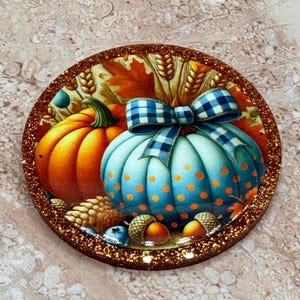 Peut inclure: Une pièce décorative circulaire présentant une scène automnale vibrante avec une citrouille orange, une citrouille bleu clair à pois orange, un nœud à carreaux bleu et blanc, des épis de blé, des glands et une bordure à paillettes cuivrées.