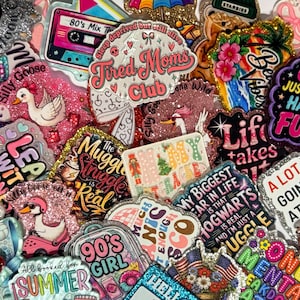 May include: A collection of assorted glittery stickers featuring various designs, including a cassette tape, a goose, text reading Tired Moms Club, Girls Just Wanna Have Fun, The Muggle Struggle Is Real, and other colorful graphic illustrations.