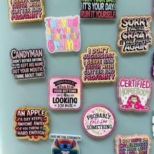 K&ouml;nnte beinhalten: Eine Sammlung bunter, glitzernder Emaille-Anstecknadeln mit verschiedenen humorvollen Phrasen. Die Anstecknadeln zeigen Texte wie "I don't sugarcoat", "Sorry I acted crazy" und "Certified Homebody". Die Anstecknadeln sind in verschiedenen Formen und Gr&ouml;&szlig;en, vor einem hellblauen Hintergrund.
