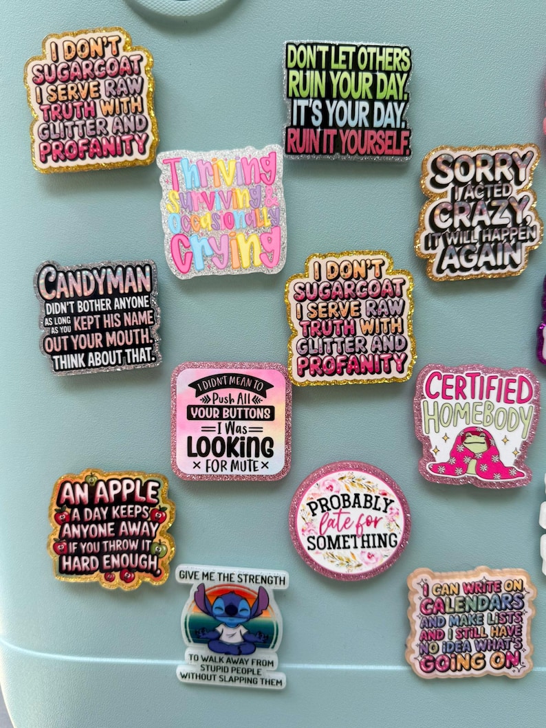 K&ouml;nnte beinhalten: Eine Sammlung bunter, glitzernder Emaille-Anstecknadeln mit verschiedenen humorvollen Phrasen. Die Anstecknadeln zeigen Texte wie "I don't sugarcoat", "Sorry I acted crazy" und "Certified Homebody". Die Anstecknadeln sind in verschiedenen Formen und Gr&ouml;&szlig;en, vor einem hellblauen Hintergrund.