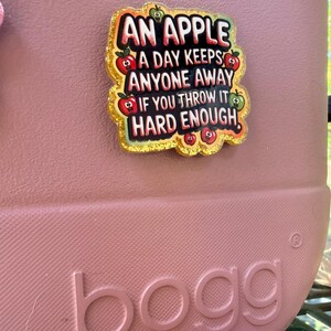 K&ouml;nnte beinhalten: Eine rosa Tragetasche mit dem Wort "bogg" auf der Seite. Eine dekorative Anstecknadel mit dem Text "AN APPLE A DAY KEEPS ANYONE AWAY IF YOU THROW IT HARD ENOUGH" ist an der Tasche befestigt. Die Anstecknadel hat einen goldenen Glitzerrahmen und Apfelgrafiken.