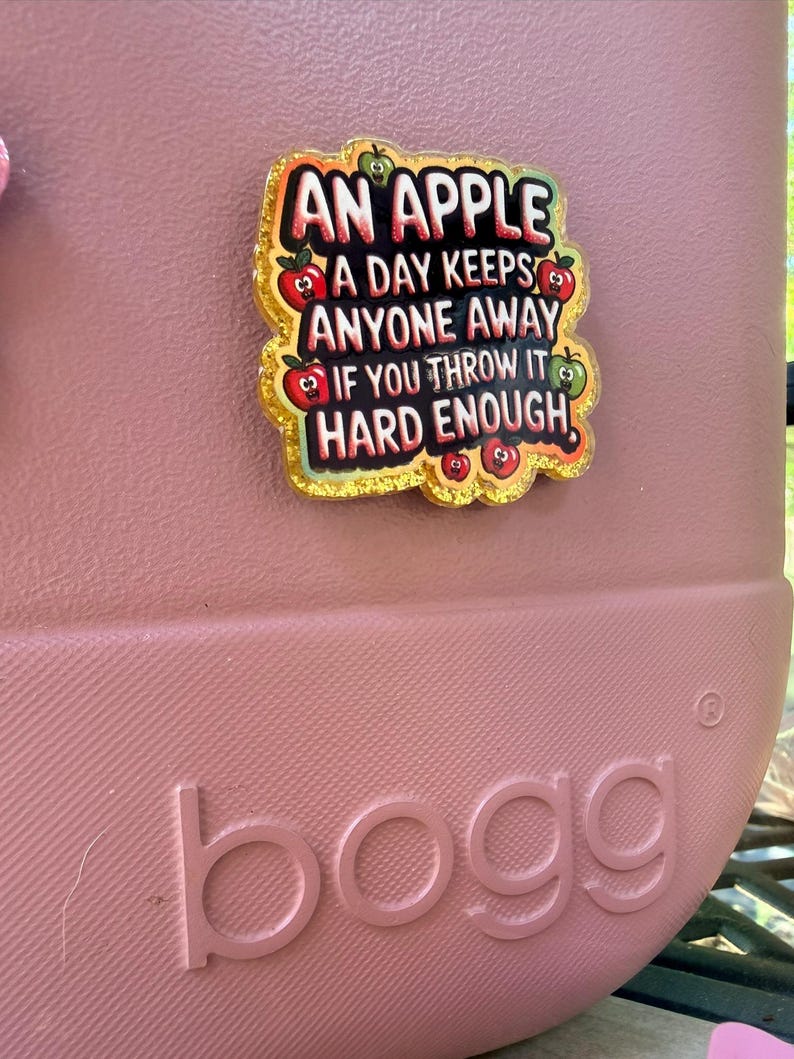 K&ouml;nnte beinhalten: Eine rosa Tragetasche mit dem Wort "bogg" auf der Seite. Eine dekorative Anstecknadel mit dem Text "AN APPLE A DAY KEEPS ANYONE AWAY IF YOU THROW IT HARD ENOUGH" ist an der Tasche befestigt. Die Anstecknadel hat einen goldenen Glitzerrahmen und Apfelgrafiken.