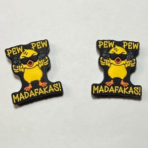 May include: Two black and yellow shoe charms. Each charm features a yellow chick wearing sunglasses and holding two guns. The text "PEW PEW" is above the chick, and "MADAFAKAS!" is below.