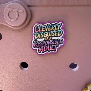 K&ouml;nnte beinhalten: Ein rosa Schuh mit einem dekorativen Anh&auml;nger mit der Aufschrift "Cleverly Disguised as a Responsible Adult". Der Anh&auml;nger hat einen glitzernden lila Rand und farbigen Text. Der Schuh hat mehrere L&ouml;cher und ein kreisf&ouml;rmiges Logo.