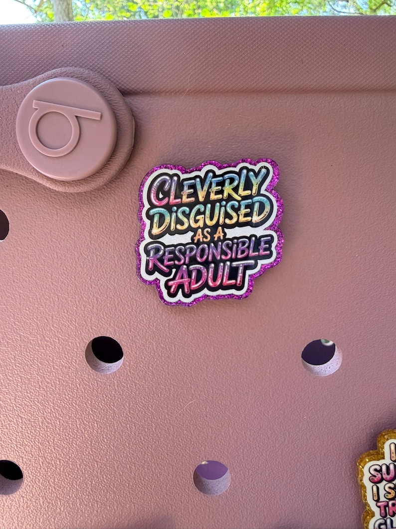 K&ouml;nnte beinhalten: Ein rosa Schuh mit einem dekorativen Anh&auml;nger mit der Aufschrift "Cleverly Disguised as a Responsible Adult". Der Anh&auml;nger hat einen glitzernden lila Rand und farbigen Text. Der Schuh hat mehrere L&ouml;cher und ein kreisf&ouml;rmiges Logo.