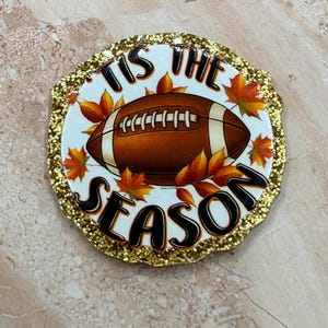 May include: A round decorative plaque with a gold glitter border featuring a brown American football in the center surrounded by orange and yellow autumn leaves. The text 'TIS THE SEASON' is printed in black lettering around the football.