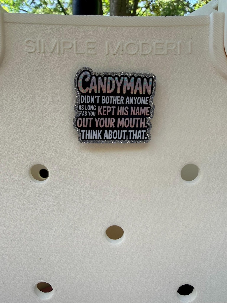 K&ouml;nnte beinhalten: Dekorative Anstecknadel mit dem Wort "CANDYMAN" und dem Zitat "DIDN'T BOTHER ANYONE AS LONG AS YOU KEPT HIS NAME OUT YOUR MOUTH. THINK ABOUT THAT." Die Anstecknadel hat einen glitzernden Rand und ist vor wei&szlig;em Hintergrund.