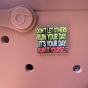 K&ouml;nnte beinhalten: Ein rosa Schuh mit einem dekorativen Anh&auml;nger mit der Aufschrift "DON'T LET OTHERS RUIN YOUR DAY. IT'S YOUR DAY. RUIN IT YOURSELF." Der Anh&auml;nger hat einen glitzernden Rand und Text in Gr&uuml;n, Gelb, Blau und Rot.