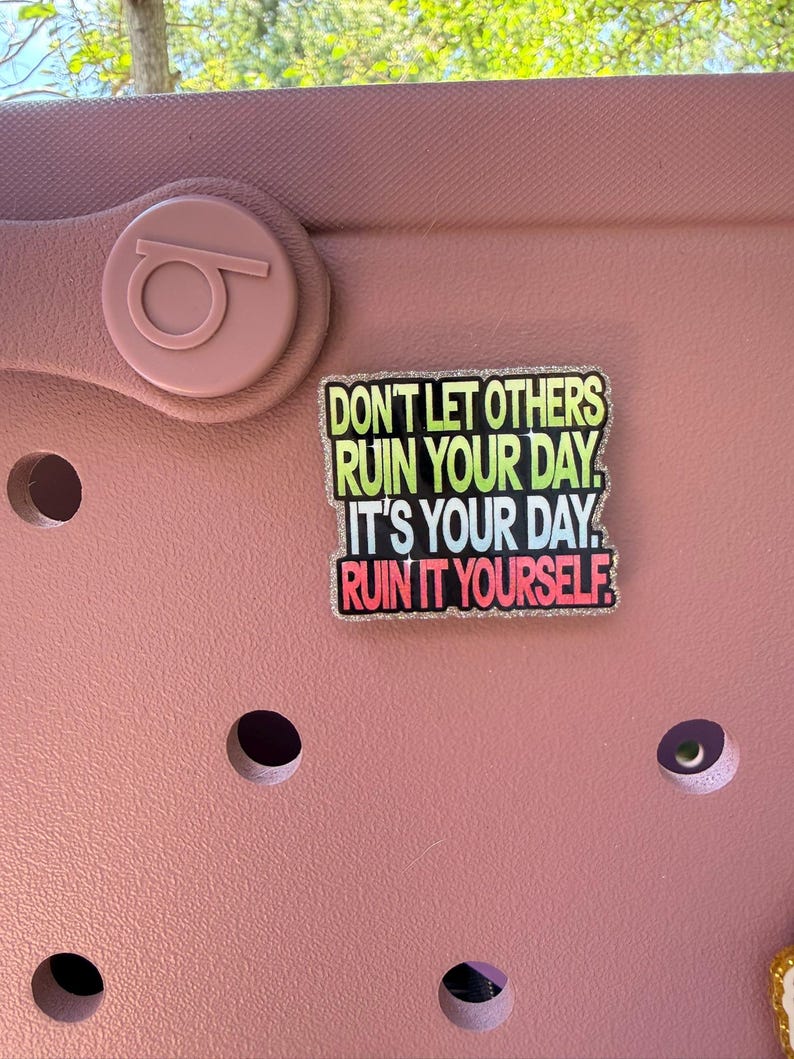 K&ouml;nnte beinhalten: Ein rosa Schuh mit einem dekorativen Anh&auml;nger mit der Aufschrift "DON'T LET OTHERS RUIN YOUR DAY. IT'S YOUR DAY. RUIN IT YOURSELF." Der Anh&auml;nger hat einen glitzernden Rand und Text in Gr&uuml;n, Gelb, Blau und Rot.