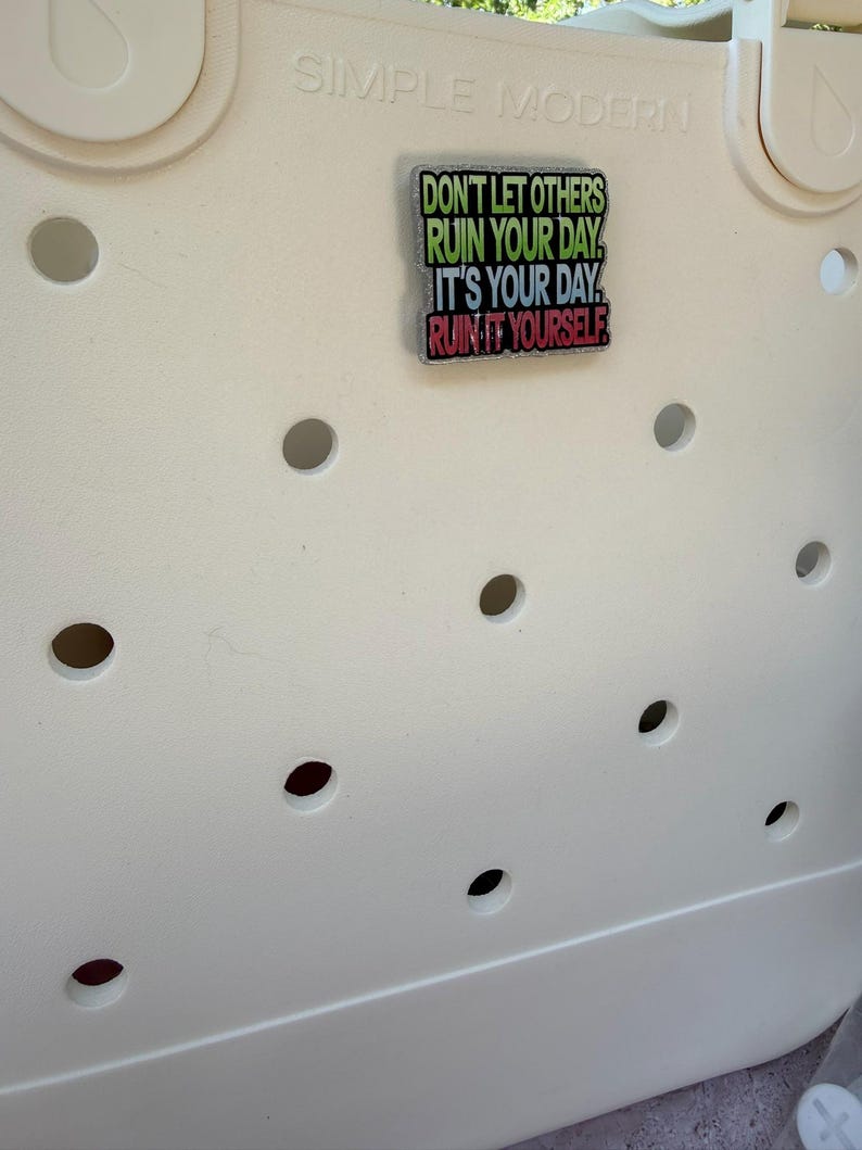 K&ouml;nnte beinhalten: Cremefarbene Tragetasche mit mehreren kreisf&ouml;rmigen L&ouml;chern. Ein rechteckiger Anstecker ist angebracht, der den Text "DON'T LET OTHERS RUIN YOUR DAY, IT'S YOUR DAY, RUIN IT YOURSELF" in Gr&uuml;n, Gelb und Rot zeigt.