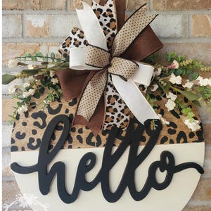 May include: Round wooden door hanger with a leopard print top and a cream-colored bottom. The word "hello" is cut out in black script. A large bow with leopard print, brown, and cream ribbons tops the sign, with greenery and white flowers.