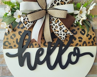 May include: A circular wooden door sign featuring a split design with a leopard print top and a solid cream bottom. The word hello is written in black script across the center. A large multi-layered bow and white flowers accent the top.