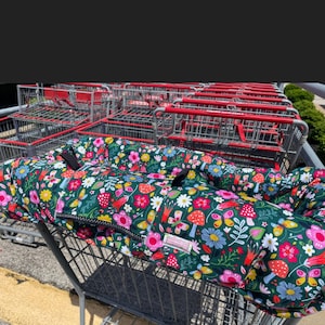 May include: A dark green shopping trolley cover with a colourful floral pattern featuring pink, white, and blue flowers, red mushrooms, and butterflies. The cover has a black zip and straps.