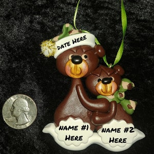 May include: A brown and green Christmas ornament featuring two bears. The larger bear is wearing a green and brown camouflage hat with the text "DATE HERE". The smaller bear is wearing a green and brown camouflage scarf. The ornament has white snow-like bases with the text "NAME #1 HERE" and "NAME #2 HERE".