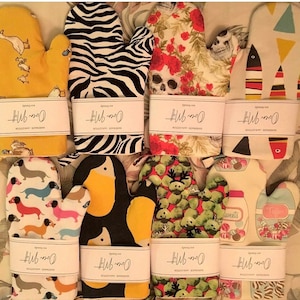 May include: A collection of oven mitts with various patterns, including a yellow mitt with ducks, a black and white zebra print, and a floral design with a skull. Each mitt has a white label with the text "Oven Mit".