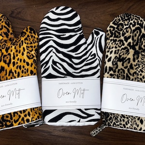 May include: Three oven mitts with animal print patterns: leopard, zebra, and leopard. Each mitt has a white label that reads "Oven Mitt" and "eco-friendly". The mitts are handmade from 100% cotton.