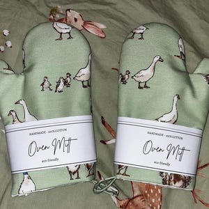 May include: A pair of green oven mitts with a white border and a white label. The label says "HANDMADE - 100% COTTON Oven Mitt eco-friendly". The mitts are decorated with a pattern of white geese and ducklings.