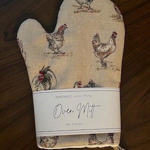 May include: Beige oven mitt featuring a repeating chicken pattern. The mitt is labelled "Oven Mitt" and "Handmade - 100% Cotton". The design is eco-friendly.