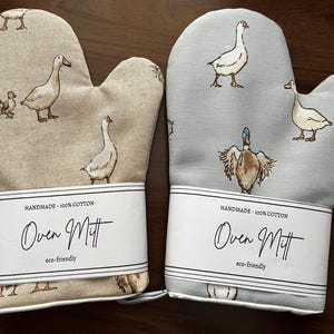 May include: Two cotton oven mitts with a goose and gosling pattern. One mitt is beige, the other is light blue. Each mitt has a white label that reads "HANDMADE - 100% COTTON" and "Oven Mitt" with "eco-friendly" below.