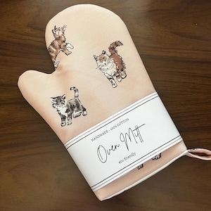 May include: A pink oven mitt with a white label that reads "Oven Mitt". The mitt features three cartoon cat illustrations in brown and grey. The label also states "handmade - 100% cotton" and "eco-friendly".