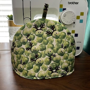 May include: A white tea cozy with a pattern of green brussels sprouts and reindeer faces. The cozy has a handle on top and is made of fabric.