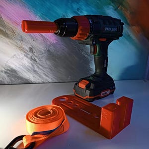 May include: A black and orange cordless drill with a red chuck and the word "PARKSIDE" printed on the side. The drill is mounted on a red plastic stand with a black and orange strap.