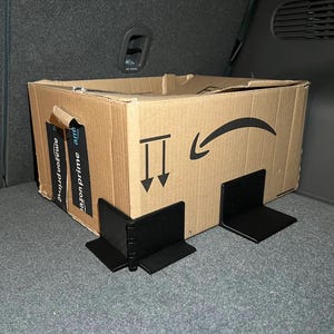 Könnte beinhalten: Ein brauner Karton mit dem Amazon-Logo und schwarzen Halterungen. Auf der Box sind ein Pfeil und zwei nach unten zeigende Pfeile aufgedruckt. Die Box befindet sich in einem Fahrzeug-Frachtraum.
