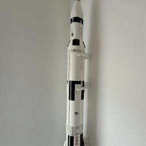 Puede incluir: Un modelo en blanco y negro del cohete Saturno V, con la palabra "USA" y una bandera estadounidense visibles. El modelo está montado en una pared blanca. El cohete tiene un diseño detallado.