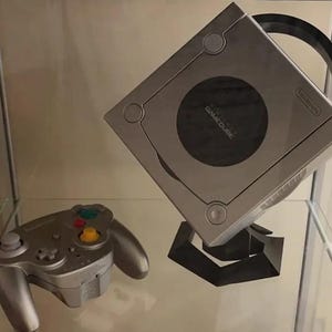 May include: A silver GameCube console with a black stand and a silver GameCube controller. The console is on a glass shelf.