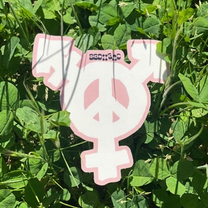 May include: A white peace sign with a pink outline, superimposed on a pink transgender symbol. The symbol is surrounded by green grass.