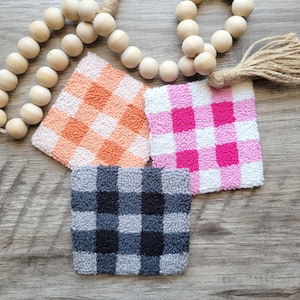 May include: Three handmade gingham-patterned coasters in orange, pink, and black and white. The coasters are made with a fluffy yarn and have a square shape.