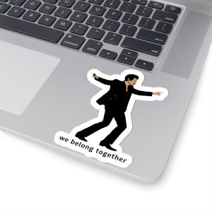 May include: A white sticker featuring a black silhouette of a person in a dynamic pose, holding a microphone and pointing. The text "we belong together" is below the figure. The sticker is placed on a laptop.