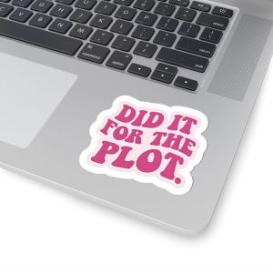 May include: A white sticker with the text "DID IT FOR THE PLOT" in pink, outlined in a lighter pink. The sticker is on a silver laptop.