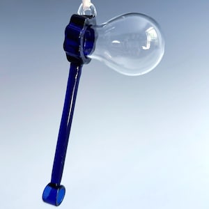 May include: A clear glass lightbulb-shaped ornament with a dark blue stem and accents. The ornament is suspended by a white string. The bulb is empty and transparent, reflecting the background. The stem is a solid, deep blue.