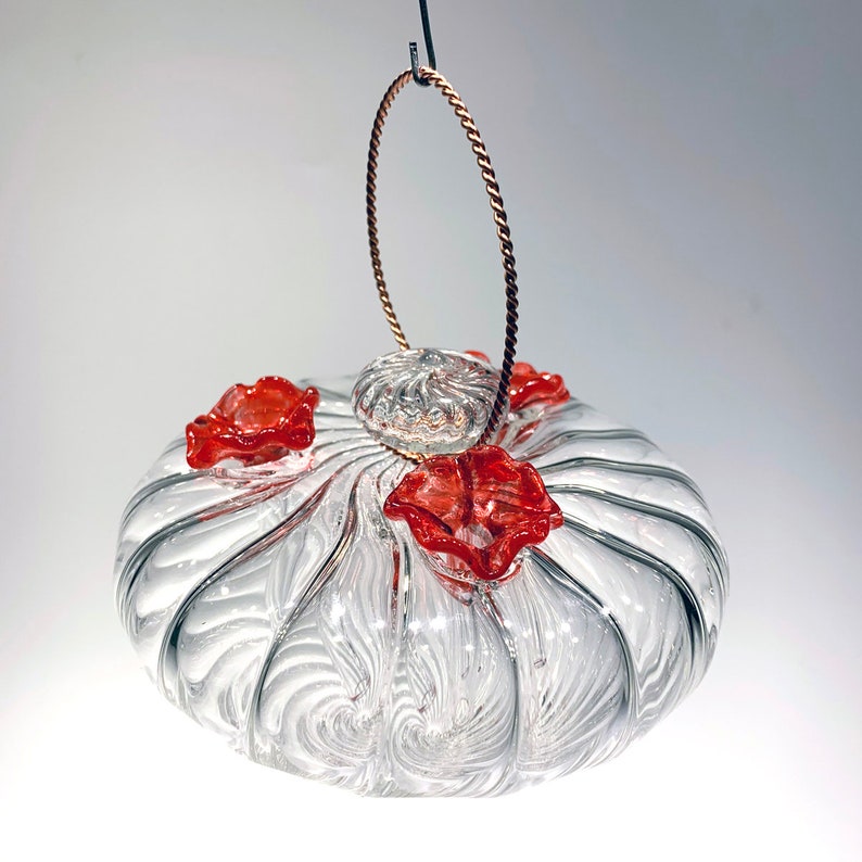 May include: A clear glass hummingbird feeder with a swirling pattern and three red flower-shaped feeding ports. The feeder hangs from a copper wire loop.