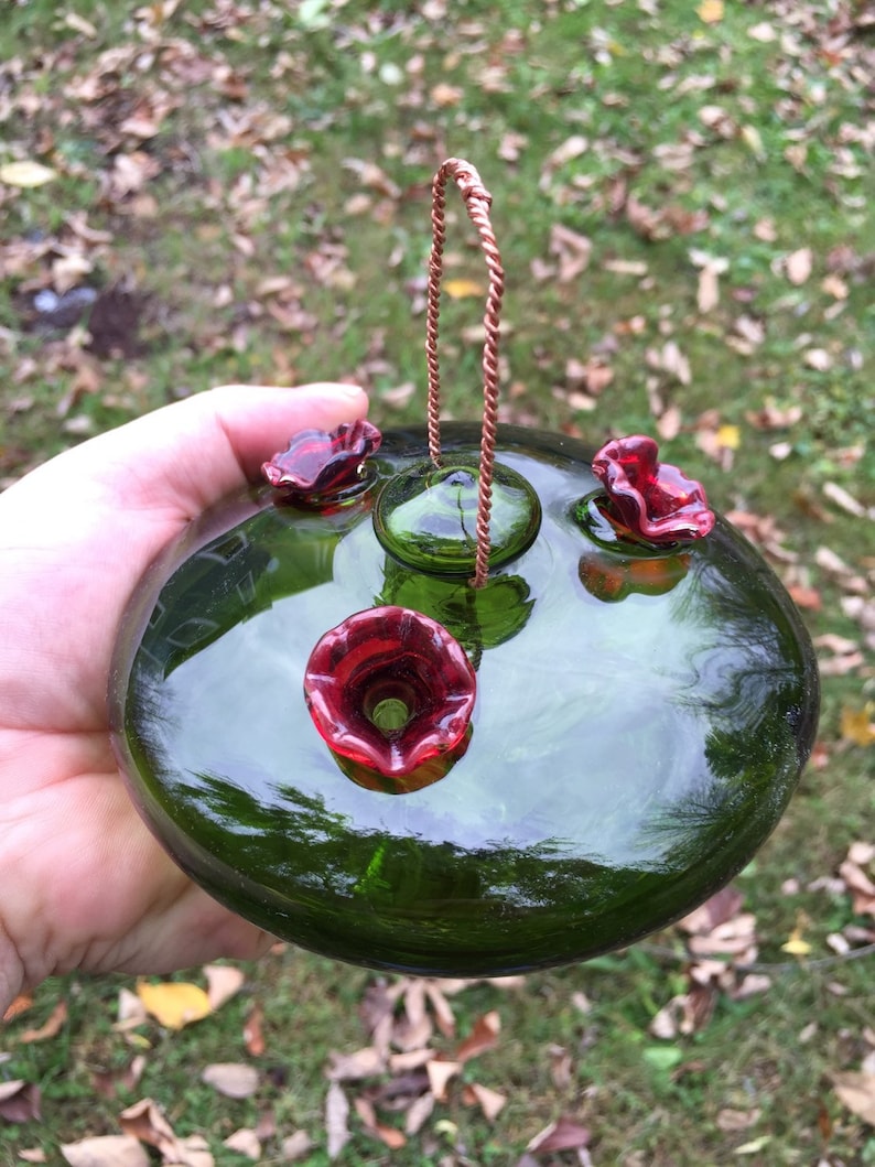 May include: A green glass hummingbird feeder with three red glass flower feeding ports and a copper wire hanger.