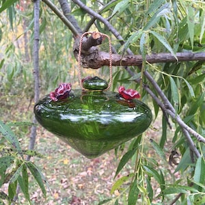 May include: A green glass hummingbird feeder with red glass flowers hanging from a tree branch.
