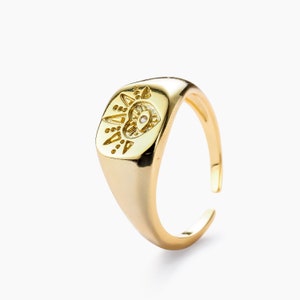 May include: A gold-colored adjustable ring with an engraved design on the face. The design includes geometric shapes and a small, round, clear stone. The ring has a polished finish and is set against a white background.