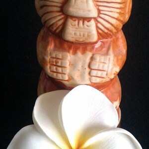 May include: A brown and tan ceramic tiki mug with a carved face and arms, paired with a white plumeria flower with a yellow center. The mug has a detailed, tribal design, and the flower is in full bloom.