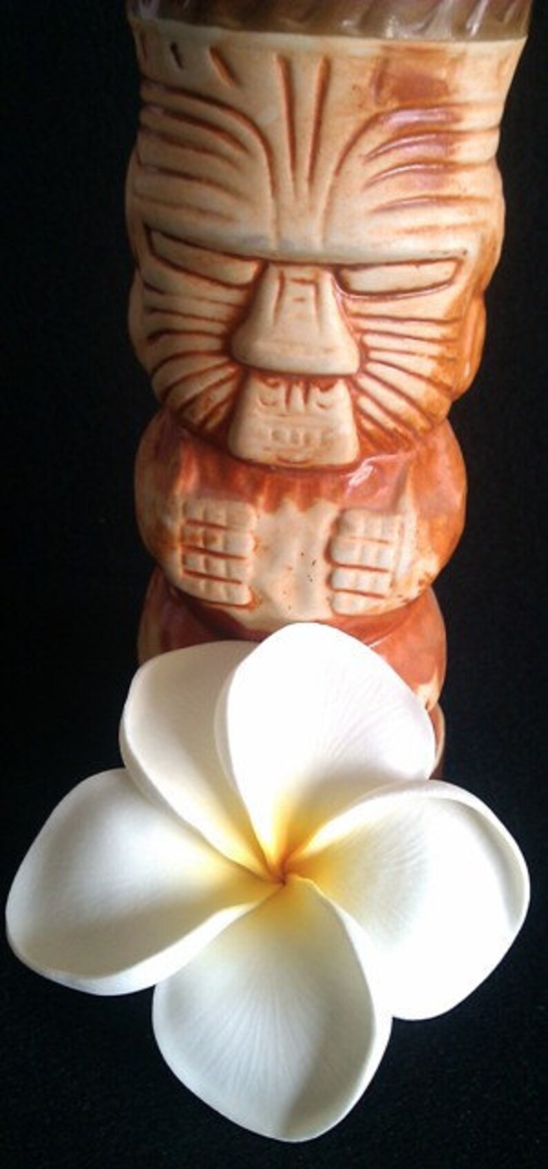 May include: A brown and tan ceramic tiki mug with a carved face and arms, paired with a white plumeria flower with a yellow center. The mug has a detailed, tribal design, and the flower is in full bloom.