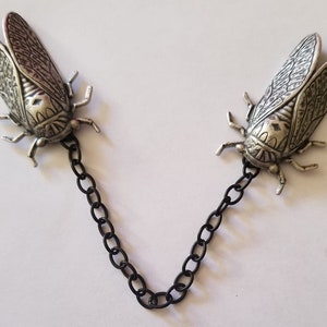 May include: A pair of silver cicada brooches connected by a black chain. The cicadas are detailed and realistic, with intricate wing patterns.