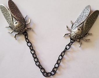 May include: A pair of silver cicada brooches connected by a black chain. The cicadas are detailed and realistic, with intricate wing patterns.