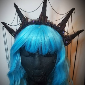May include: A black crown with dark purple crystals and chain accents sits atop a black mannequin head with bright blue hair. The crown has multiple points and a gothic aesthetic, suitable for cosplay or themed events.