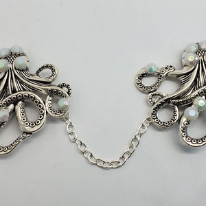 May include: A pair of silver-toned octopus-shaped brooches connected by a chain. Each octopus has multiple tentacles and is adorned with iridescent, pearlescent beads. The brooches are set against a white background.